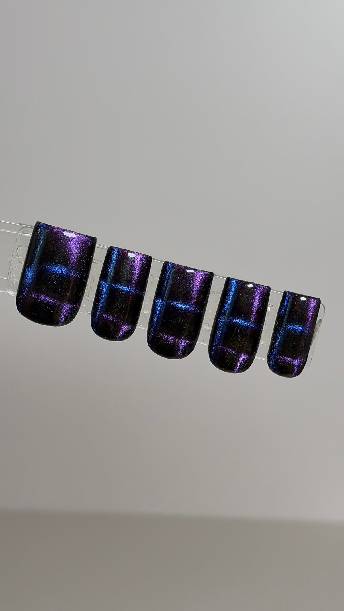 Five false press-on nails with a dark purple and black base, decorated with shimmering blue and purple cat-eye magnetic stripes, shown on a clear display stand against a neutral light gray background.
