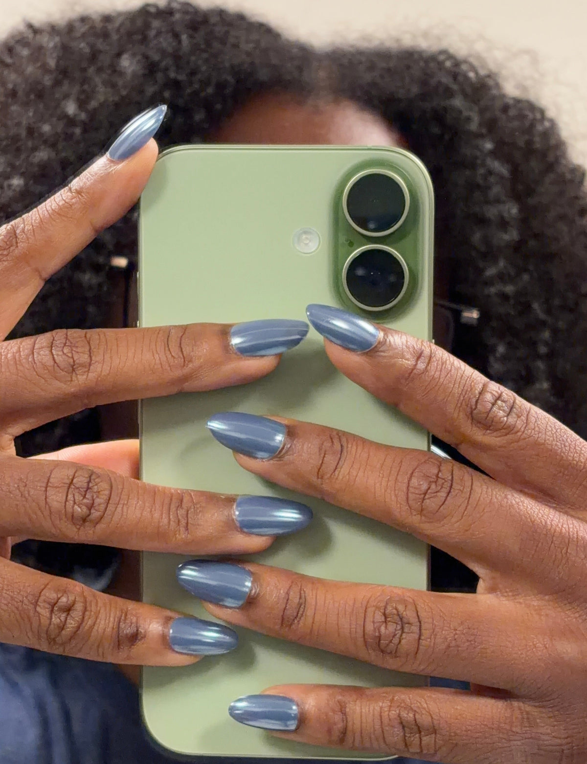 A close-up mirror selfie showing a person with dark curly hair holding a light green smartphone. The person is wearing a blue-gray metallic nail polish on all their nails.
