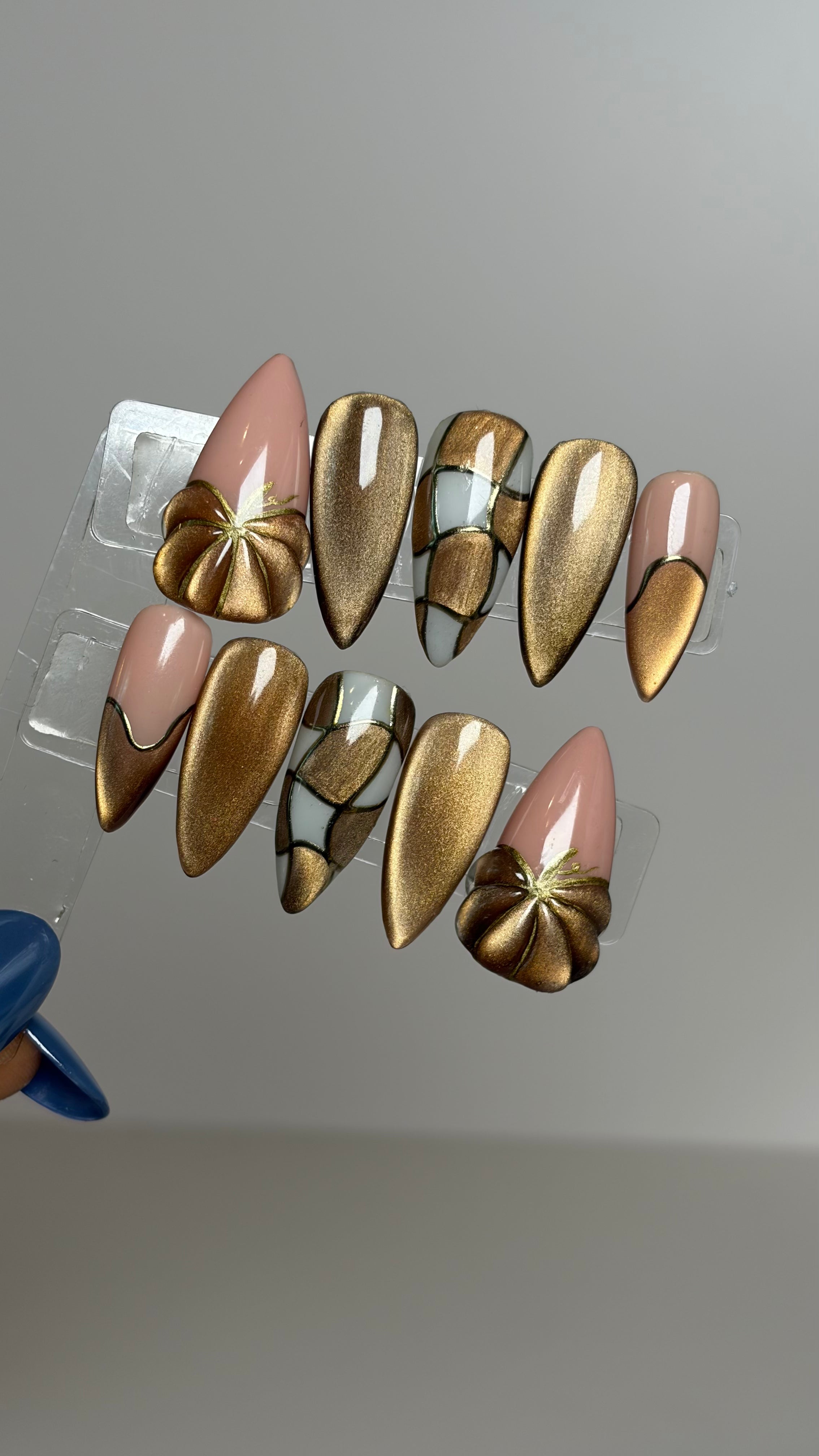 A set of decorative press-on nails displayed on a clear plastic hanger against a plain light gray background. The nails feature metallic gold and champagne-gold gradient designs, some with glossy nude pink tips and others with metallic rose-gold and gold floral patterns.