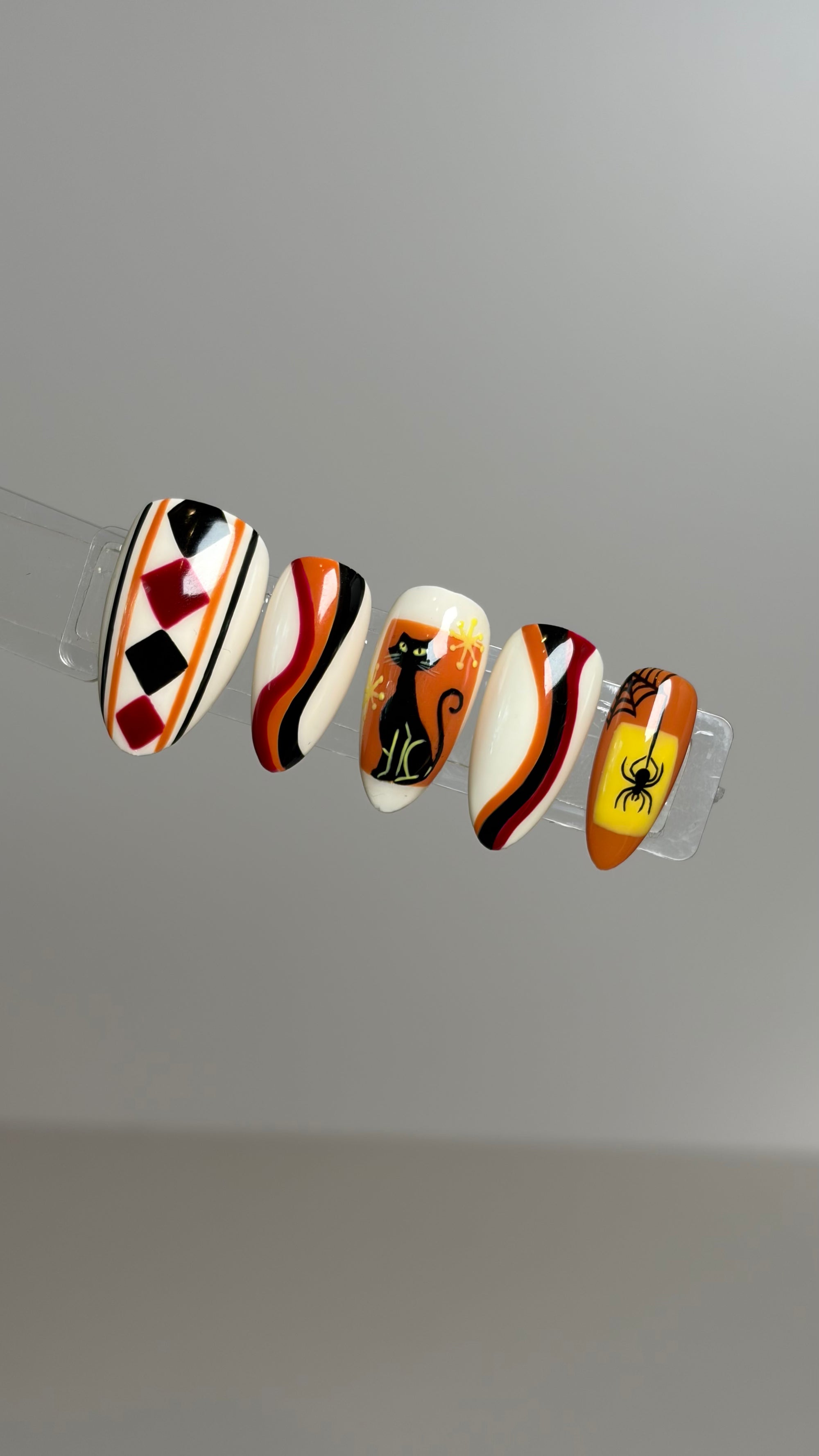 Five decorative false nails displayed on a clear stand against a plain light gray background. The nails feature Halloween-themed designs including a harlequin pattern, stripes, a black cat silhouette, a pumpkin with a sun, and a spider on an orange background.