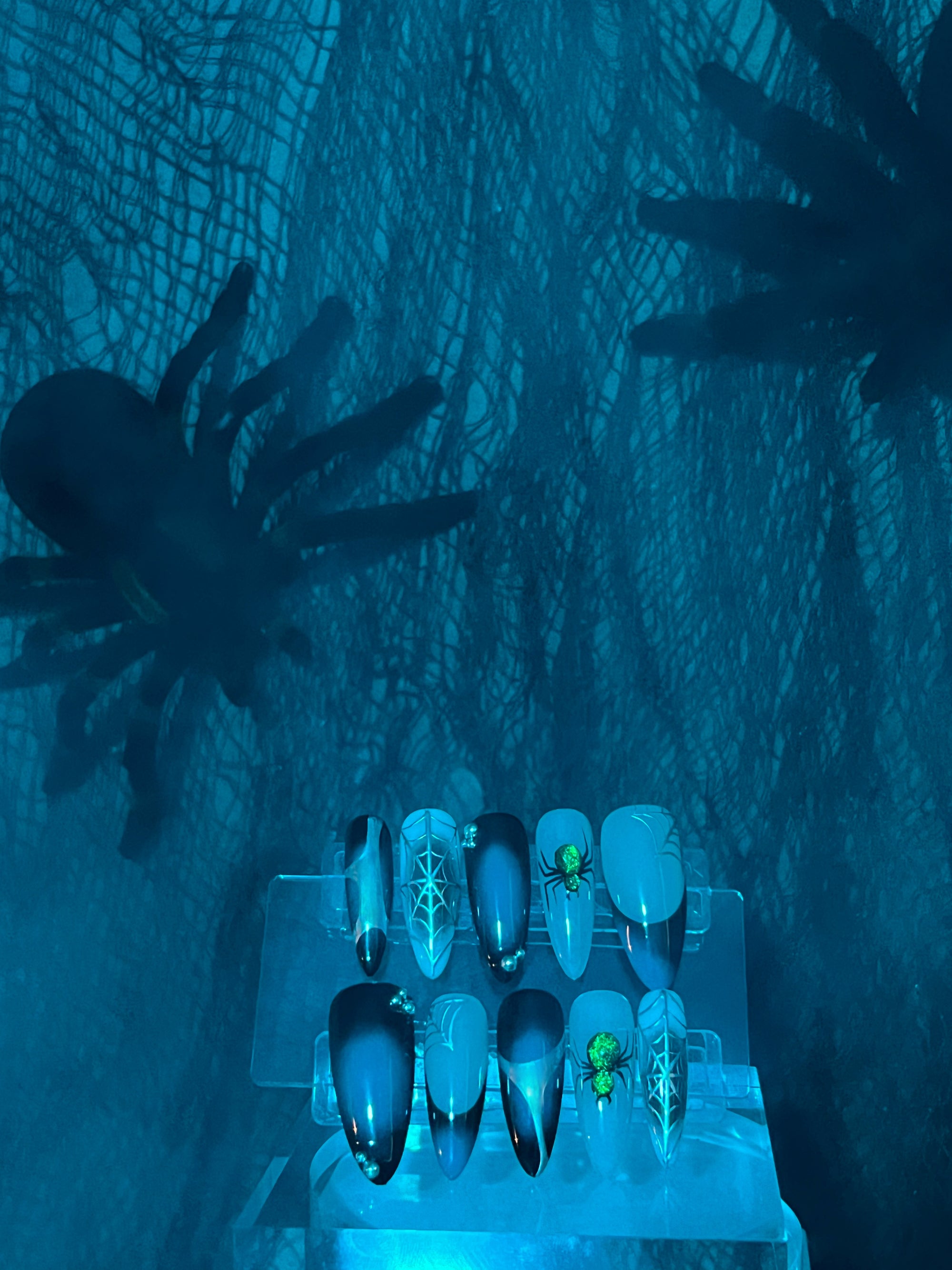 A set of press-on nails with 3D spider web details, displayed on a blue surface with spider decorations in the background, under a blue light.