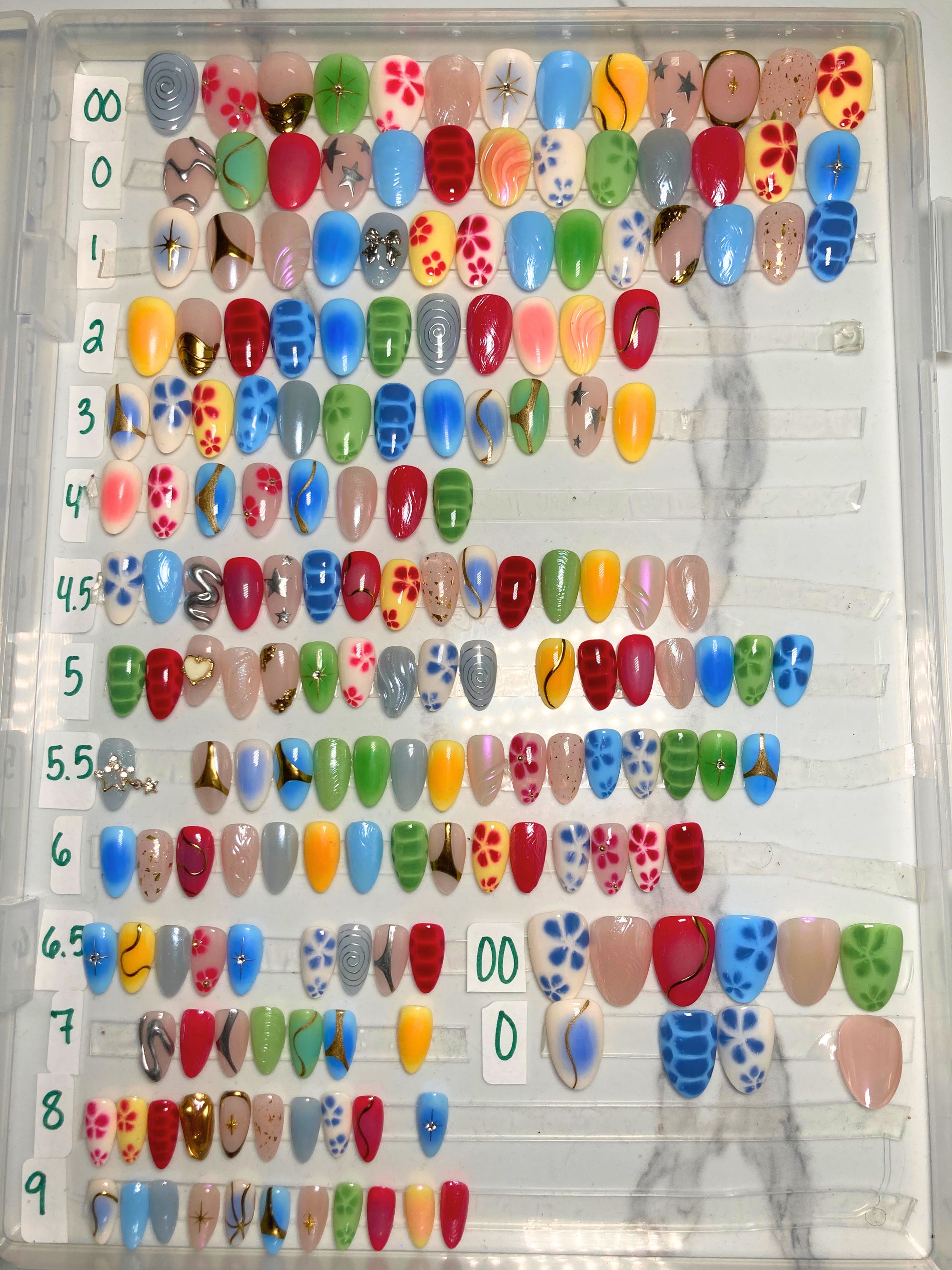A clear plastic tray displaying multiple rows of artificial press-on nails in assorted colors and patterns, including solid shades, polka dots, stripes, floral motifs, and metallic finishes.