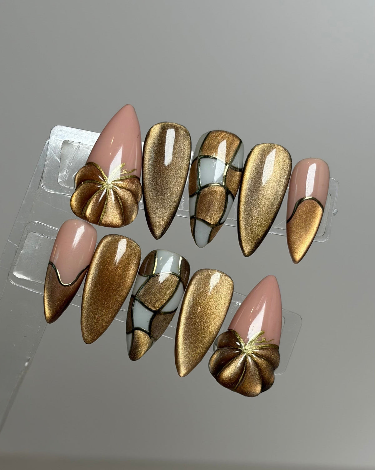 A set of artificial press-on nails displayed on a clear plastic stand against a neutral gray background. The nails feature a mix of metallic gold cat eye designs, solid pale pink tips, and some with silver chrome accents.